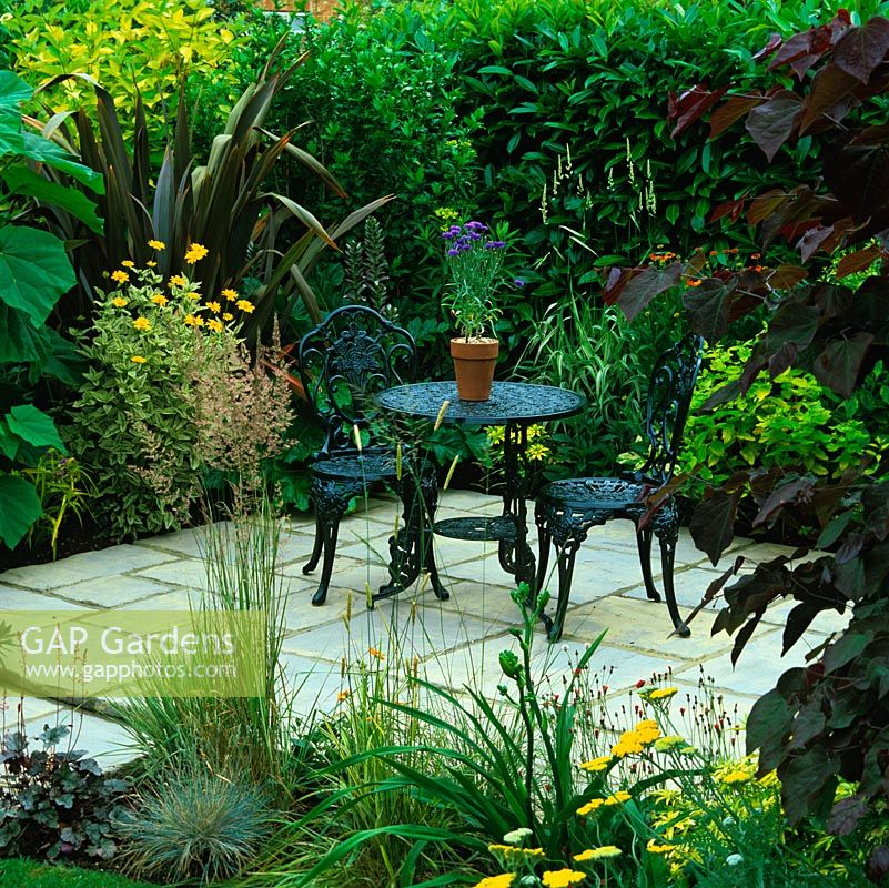 Dans un petit jardin de banlieue, une cour secrète avec table et chaises, cachée derrière des cercis, des tulipiers, du phormium, des herbes et des haies de lauriers.