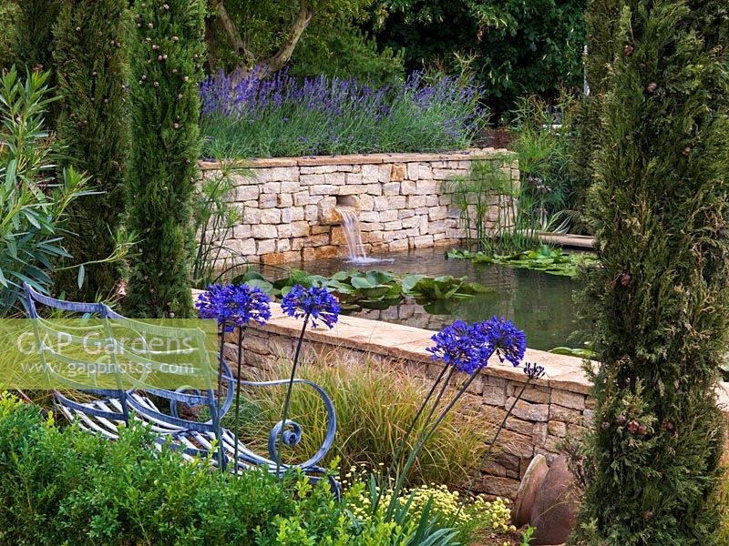 La piscine surélevée, recouverte de murs en pierre sèche, abrite des nénuphars. Jardin de style méditerranéen avec lavande, agapanthe, cyprès, olive, laurier-rose et santoline. Banc.