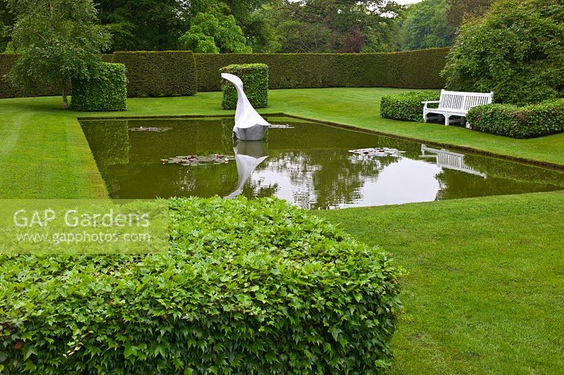 Un étang de nénuphars tranquilles avec une sculpture de baleine surplombée par un siège et entouré de pelouse, topiaire et une haie de Taxus à Farleigh House, Hampshire