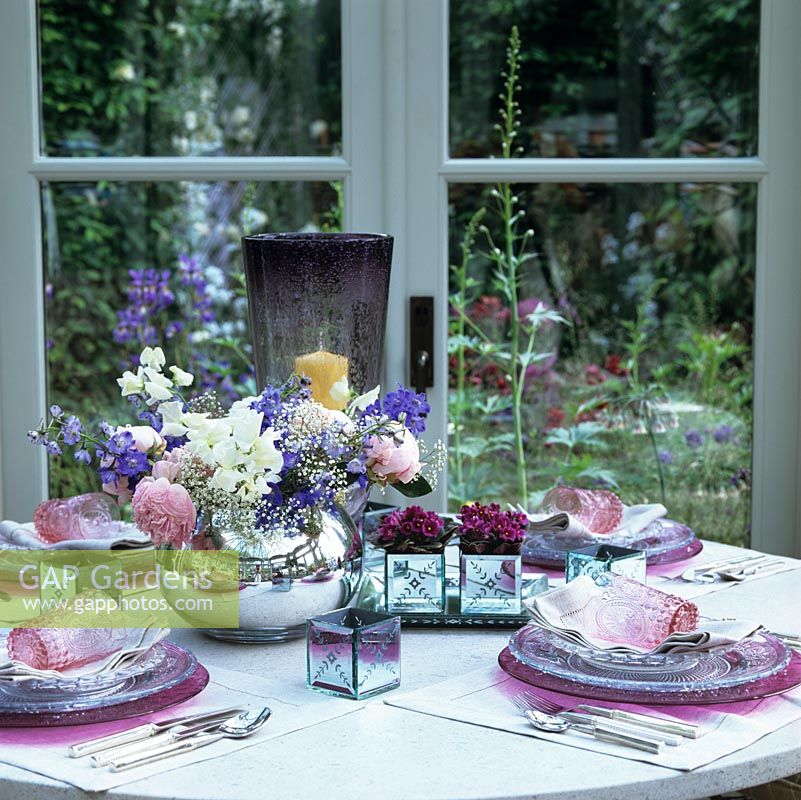 Joli vase de petits pois, de roses, de delphinium et de gypsophile au centre d'une table joliment dressée dans une véranda. Vues du jardin au-delà.