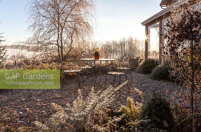 Véranda, terrasse en gravier avec mobilier de jardin en métal et parterres de fleurs mixtes avec saule pleureur en arrière-plan - décembre, Mas de Bety, France