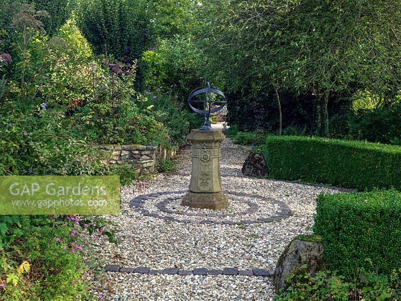 Un cadran solaire ornemental au centre d'un parterre de gravier rond dans le jardin en terrasse.