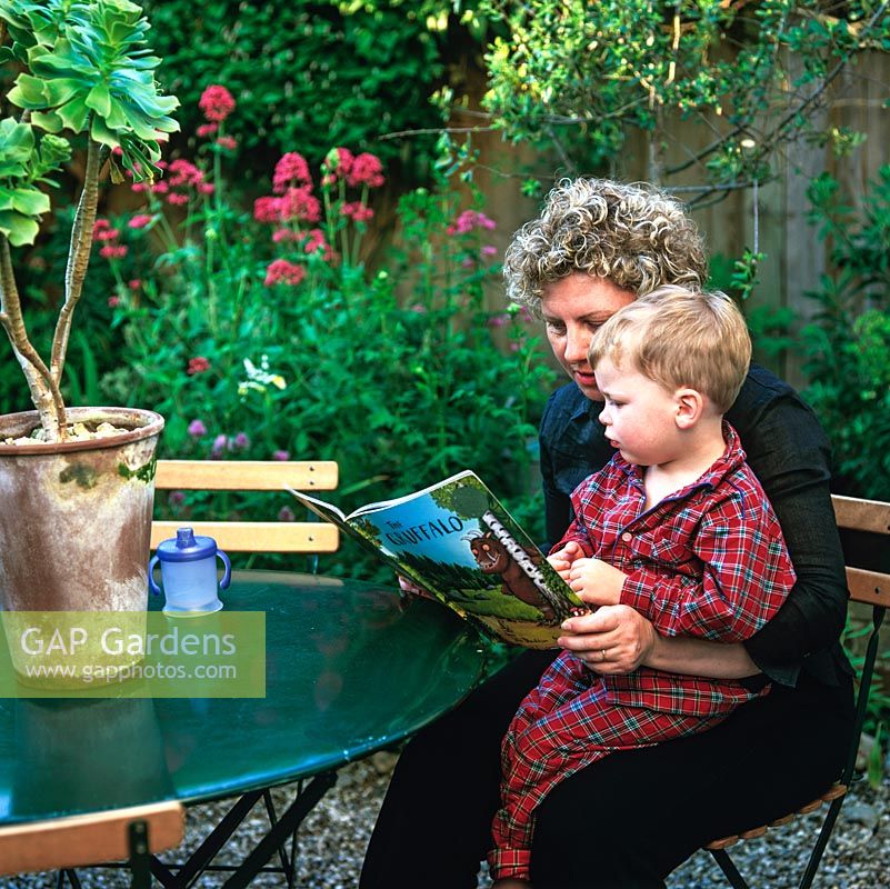 Mhairi Clutson lisant une histoire de parterre de fleurs à son plus jeune enfant, Phelin, dans leur petit jardin à l'arrière de style méditerranéen avec son patio en gravier en contrebas au-dessous d'une cour fermée.