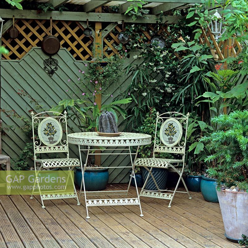 Chaise et tables sur terrasse en bois ombragée, cachées derrière des panneaux de clôture, des pots et des jasmoïdes Trachelospermum, nichées au fond d'un jardin de ville de 27 mx 13 m.