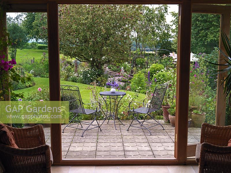 Les portes-fenêtres donnent sur la terrasse et le jardin de campagne au-delà avec des parterres de lys, de roses, de cosmos, de Verbena bonariensis, de lupin, d'allium, de buddleja et de vieux pommier.