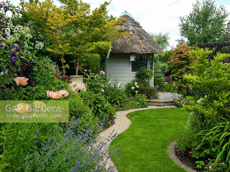 Un chemin d'herbe en forme mène à travers des parterres mixtes matures à une maison d'été au toit de chaume.