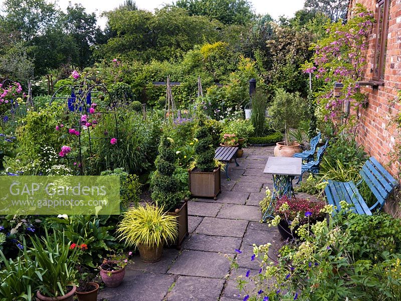 Terrasse en pierre avec des sièges et une collection de pots et de spirales topiaires donnant sur le jardin parterre.