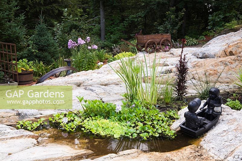 Sculpture de jeune garçon et fille en barque dans un étang dans la cavité rocheuse avec Eichornia crassipes - Jacinthe d'eau, Typha latifolia - Plantes de quenouilles communes, fleurs Phlox violettes