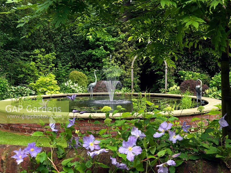 Entrevu à travers des clématites sur un mur de jardin, une piscine circulaire surélevée avec fontaine centrale.