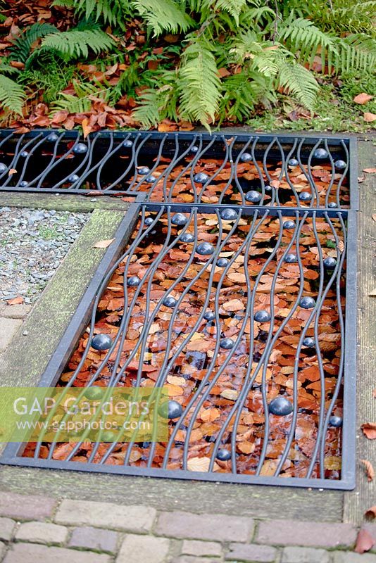 Petit étang carré rempli de feuilles d'automne brunes flottantes recouvertes d'une grille métallique décorée de noir dans le trottoir.