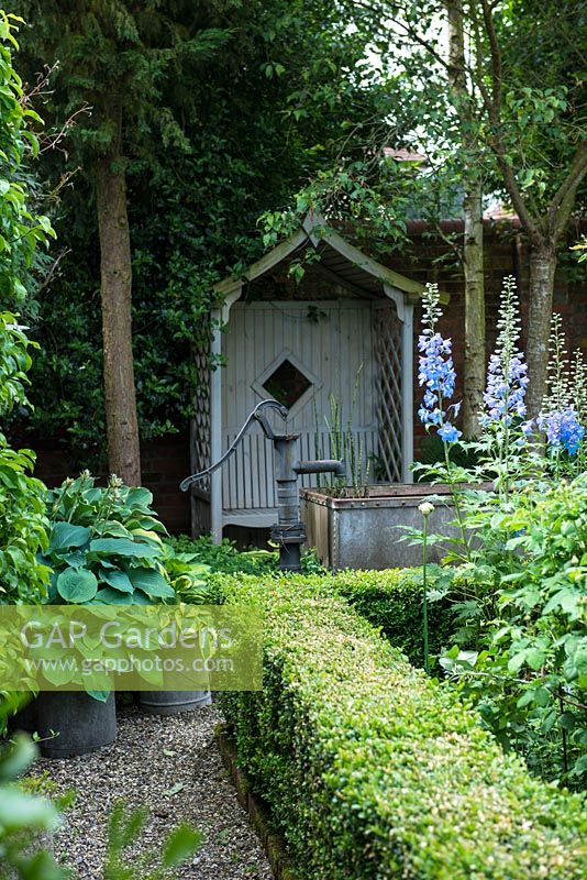Un coin ombragé d'un jardin clos avec un siège en bois couvert, un petit étang fait d'un abreuvoir en métal galvanisé et d'une plantation tolérante à l'ombre, y compris Hosta et des haies de buis.
