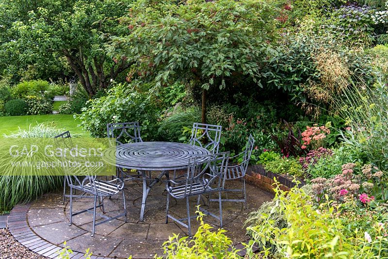 Un patio circulaire en pierre avec table et chaises. La plantation dans le parterre de fleurs surélevé environnant comprend Sedum, Achillea, Stipa gigantea, Viburnum et un arbre Sorbus.