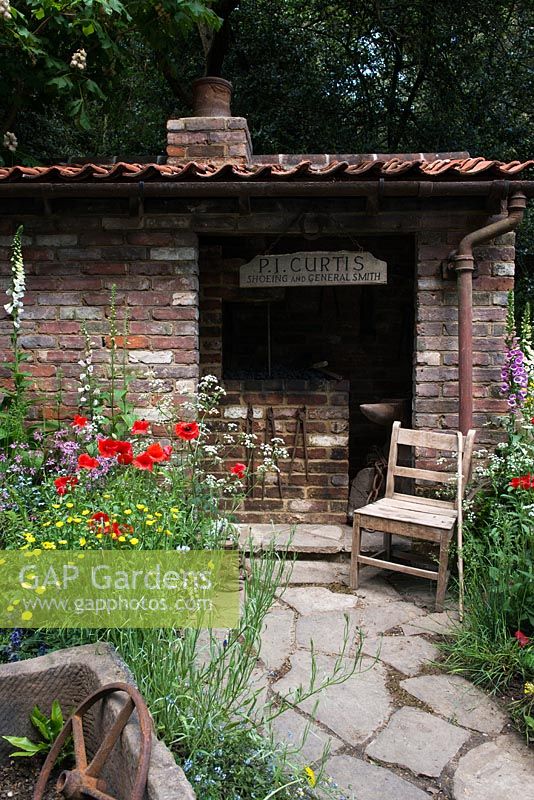 The Old Forge Artisan Garden for Motor Neurone Disease Association - RHS Chelsea Flower Show 2015. Vue des sentiers de roche naturelle et un hangar en brique et une chaise vintage entourée de fleurs sauvages