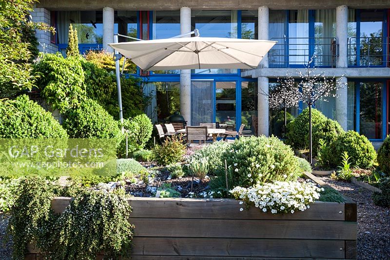 Maison et terrasse avec des sièges, un éclairage de jardin, un parasol et un parterre d'éboulis surélevé entouré de topiaires - mai, Scalabrin Laube Garten, Suisse