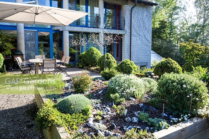 Maison et terrasse avec des sièges, un éclairage de jardin, un parasol et un parterre d'éboulis surélevé entouré de topiaires - mai, Scalabrin Laube Garten, Suisse