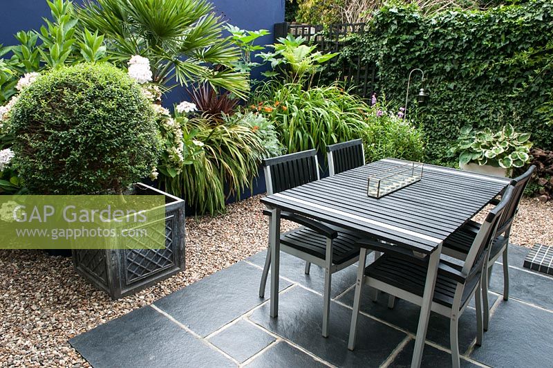 Une salle de jardin conçue pour recevoir avec un pavé gris anthracite surmonté d'une table et de chaises, un mur de cobalt, une clôture recouverte de lierre et des feuilles audacieuses de palmiers et de cordylines.