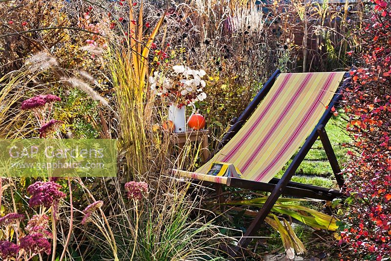 Chaise de terrasse en bois parmi les vivaces et les graminées en automne. Pennisetum, Crocosmia 'Lucifer', Aster, Sedum, Panicum virgatum.