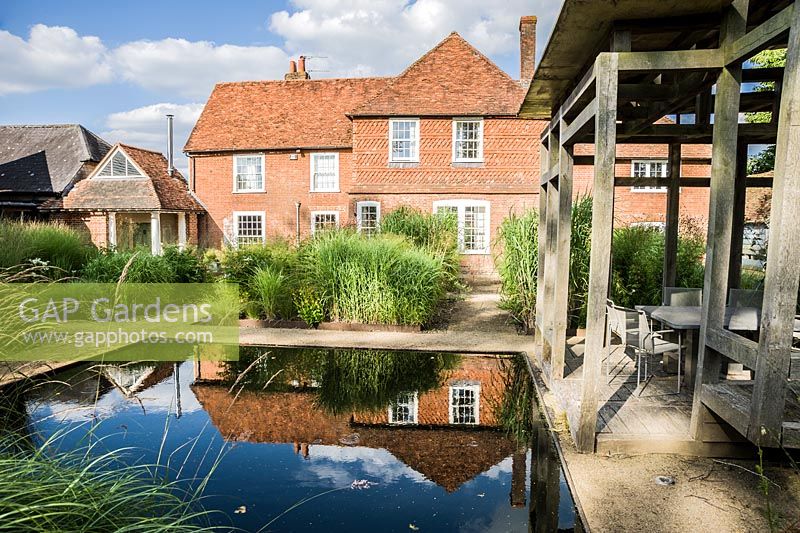 Au centre du jardin avant minimaliste, conçu par Christopher Bradley-Hole, se trouve une maison d'été contemporaine en chêne vert à côté d'une piscine immobile entourée de miscanthus et d'autres herbes hautes. Bury Court Barn, Bentley, Hants, Royaume-Uni