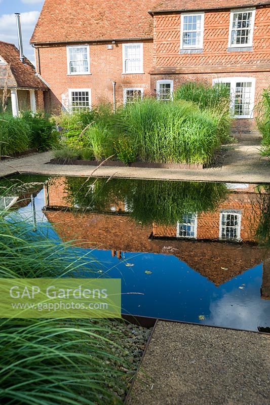 Au centre du jardin avant minimaliste, conçu par Christopher Bradley-Hole, se trouve une piscine immobile entourée de grands miscanthus et d'autres herbes. Bury Court Barn, Bentley, Hants, Royaume-Uni