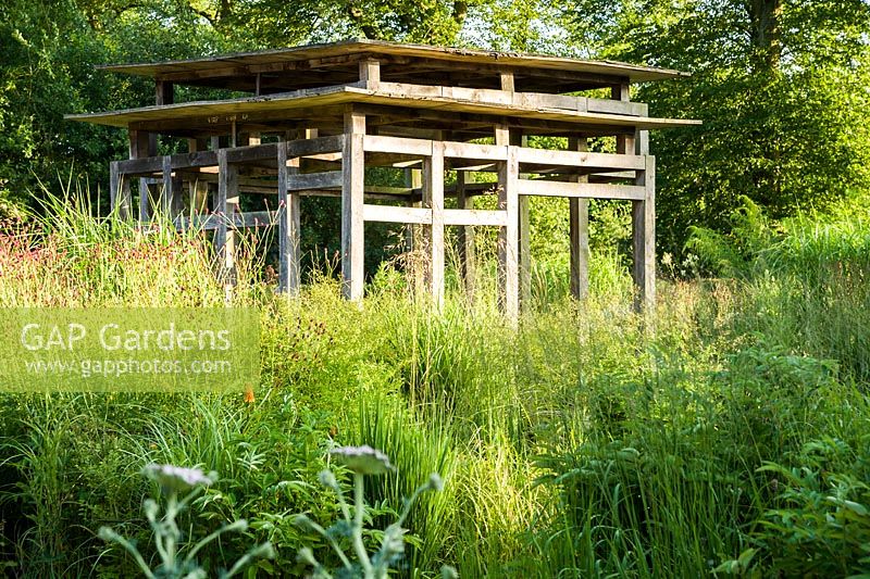 Au centre du jardin avant minimaliste, conçu par Christopher Bradley-Hole, se trouve un pavillon d'été contemporain en chêne vert entouré de hauts miscanthus et autres herbes mélangées à des plantes vivaces herbacées, y compris des sanguisorbas. Bury Court Barn, Bentley, Hants, Royaume-Uni