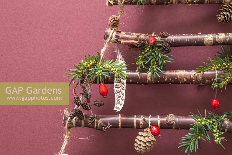 Un arbre de Noël suspendu fait avec des branches de bouleau, sur un fond rouge. Les décorations comportent des pommes de pin, des plumes, des cynorrhodons et du feuillage de pin