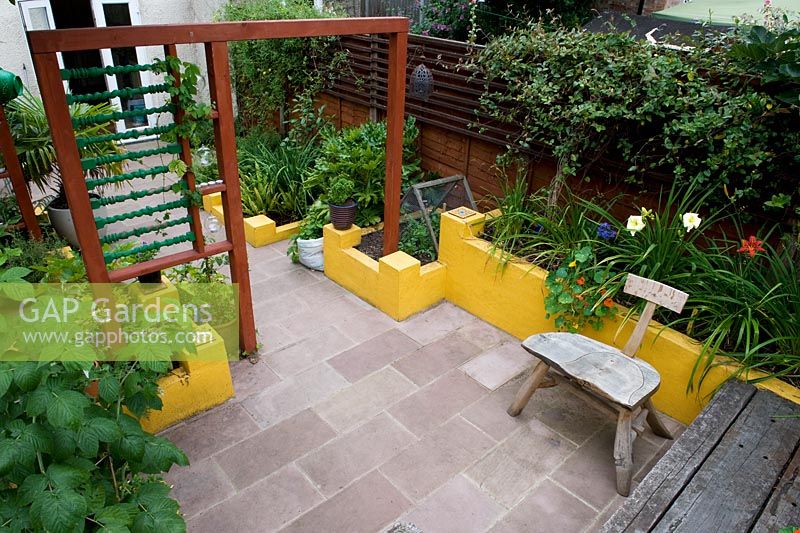 Vue d'en-haut. Sol en pierre d'aménagement paysager, murs bas peints en jaune, séparateur de jardin en bois créant deux pièces et plantation de feuillage vert.