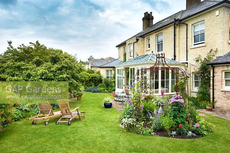 Vue sur jardin familial informel avec chaises longues sur la pelouse, tonnelle en métal, parterres de fleurs mixtes et véranda.