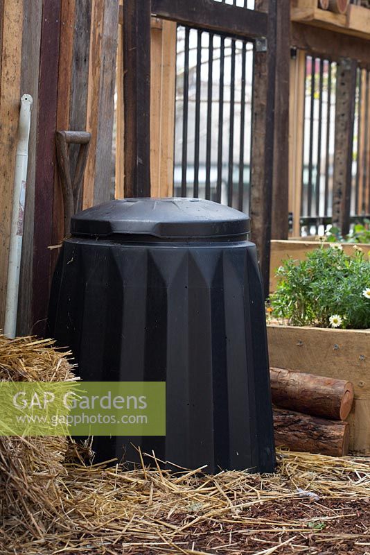 Un bac à compost en plastique noir autoportant contre un mur en bois dans un jardin communautaire.