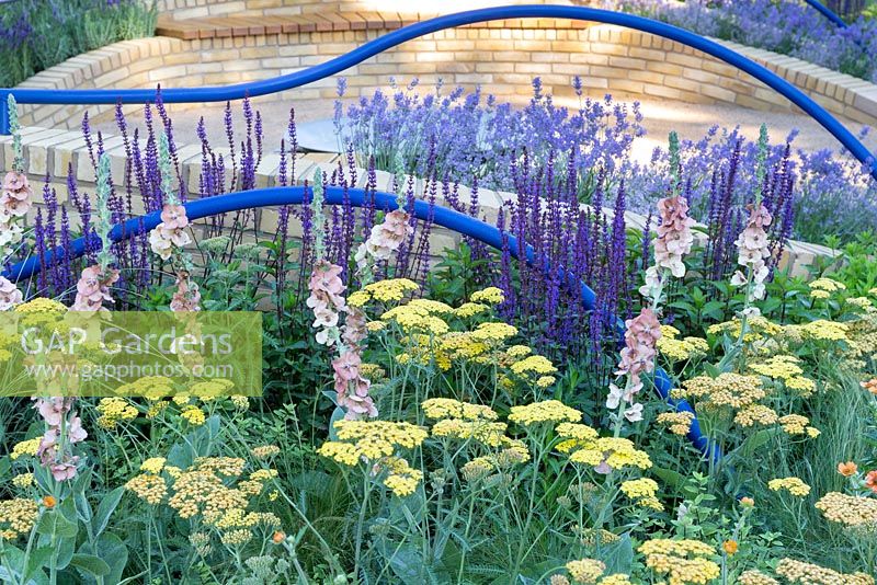 A Breath of Fresh Air, le jardin visait à créer un espace apaisant pour les personnes atteintes de démence. Des mains courantes incurvées bleu vif se trouvent au-dessus des bancs de briques dans une zone fermée avec une pièce d'eau circulaire en métal au centre. Les plantes comprennent l'achillée, le verbascum, le geum et la salvia. RHS Hampton Court Flower Show en 2016