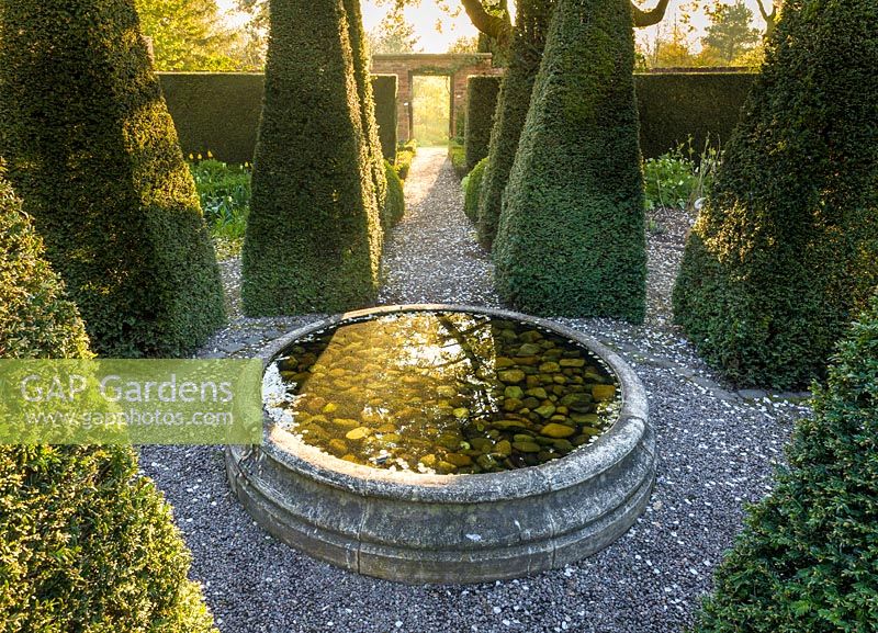 La lumière de l'aube brille à travers une porte d'entrée dans le jardin du puits avec ses ifs pyramidaux et ses tulipes, au Wollerton Old Hall Garden, Shropshire - photographié en avril
