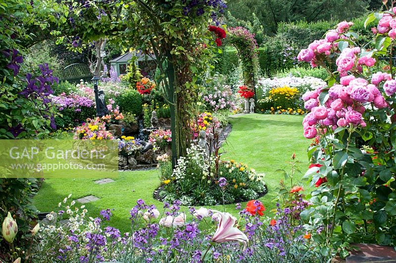 Vue sur petit jardin arrière coloré avec des parterres remplis de plantes annuelles tendres, vivaces, Rosa 'Pomponella' et Clematis sur treillis et étang