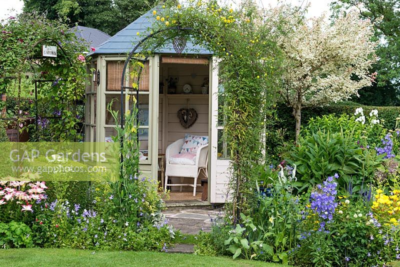 Un jardin de chalet avec maison d'été, arc couvert de clématites, boîte topiaire et parterre de fleurs mélangé coloré