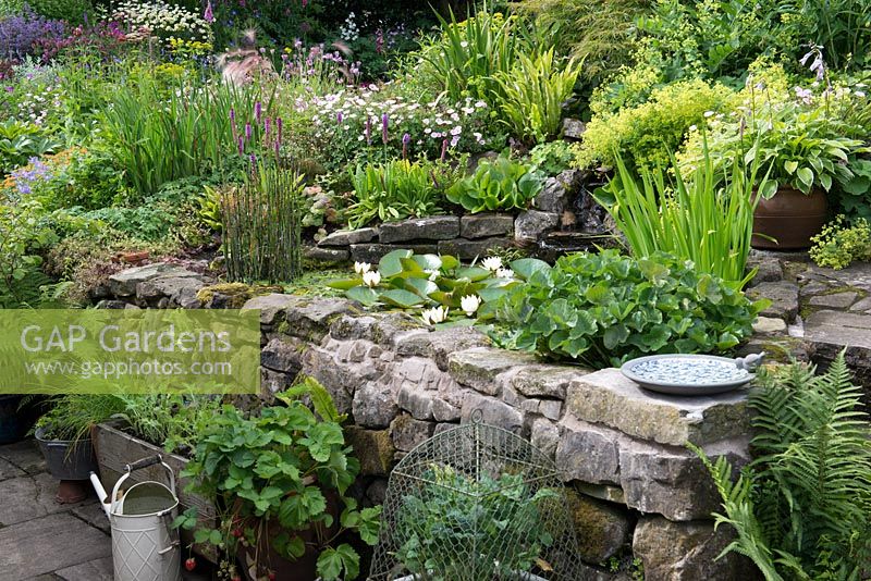 Un jardin de chalet avec étang surélevé, jardin en pot cultivant des fruits, des herbes et des légumes et un grand parterre de fleurs en pente avec des vivaces mixtes.