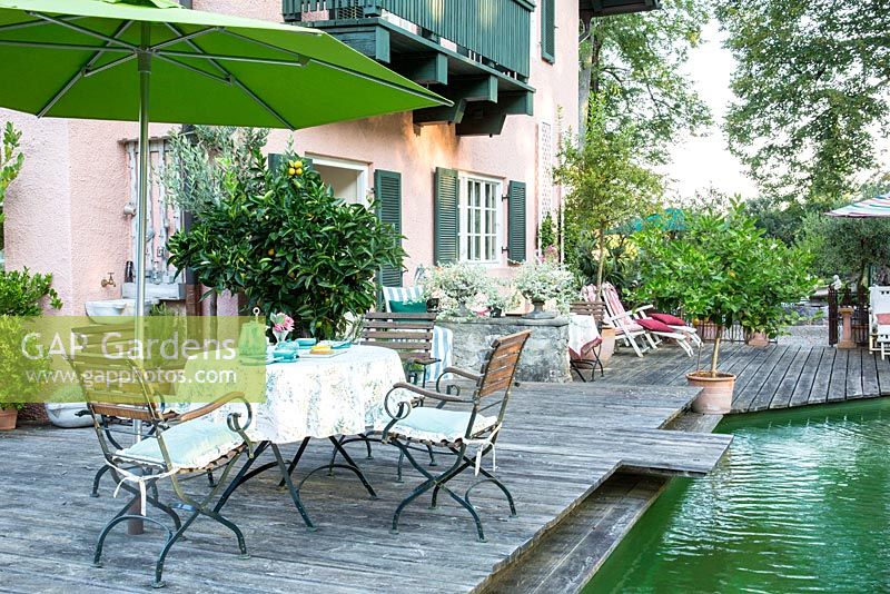 Sur la terrasse en bois entre une maison et un étang de baignade, des meubles de jardin en bois, un muret en pierre et des plantes en pots, dont des agrumes