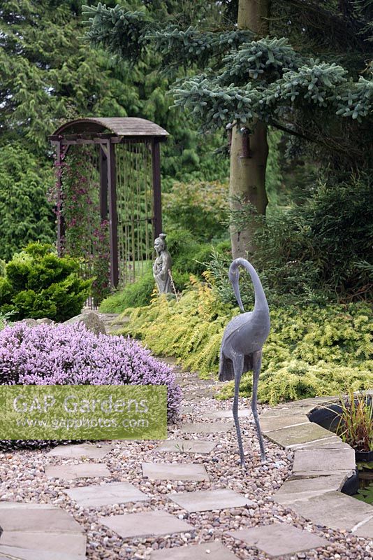 Une sculpture de héron en métal au bord d'un étang avec une arche et des arbres au-delà dans un jardin de style japonais. Juin, North Yorkshire.