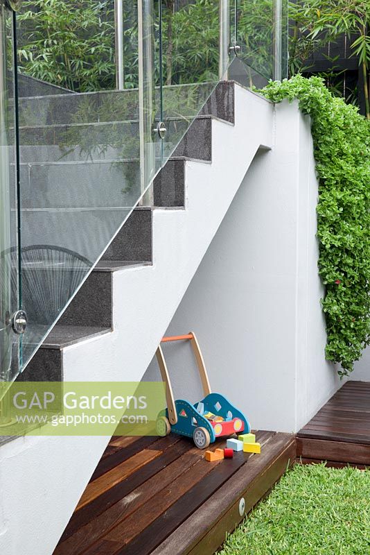 Une aire de jeux pour enfants sous un escalier avec des écrans de verre sans cadre, une terrasse en bois, un jouet pour enfants en bois et Aptenia cordifolia, une banquise au cœur qui déborde sur un mur enduit de ciment peint en gris.