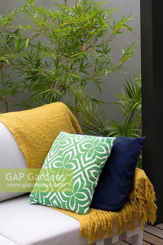 Détail d'un salon extérieur avec un tapis jaune, à motifs géométriques verts et blancs et un coussin bleu marine devant un Bambusa textilis 'Gracilis', des tisserands élancés en bambou et un mur de cour enduit de ciment.
