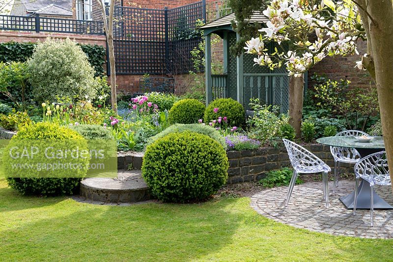 Un jardin clos de ville avec un coin salon sur un patio circulaire, des parterres de fleurs mixtes surélevés avec une boîte, des hébreux et des tulipes et un kiosque en bois peint.