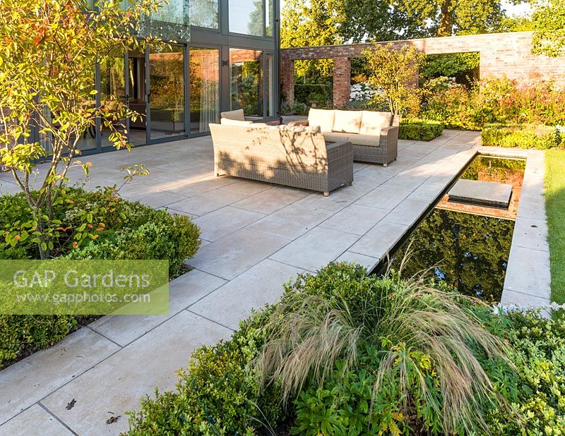 Un patio, avec des sièges et une rigole, dans un jardin de campagne moderne du Cheshire, conçu par Louise Harrison-Holland. La plantation comprend Amelanchier lamarckii, la couverture de boîte et Stipa tenuissima