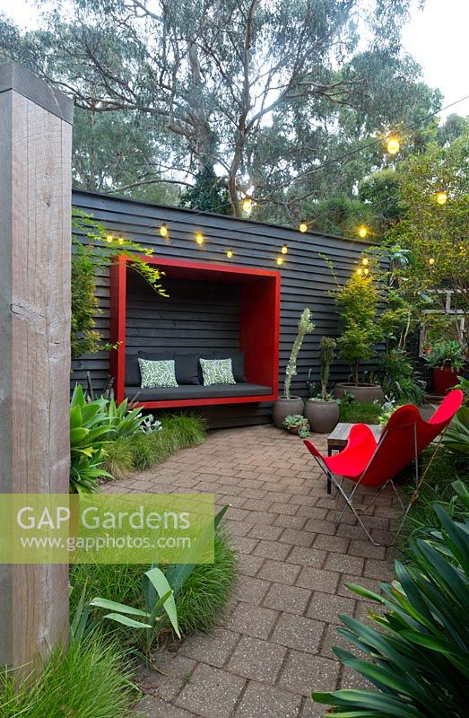 Zone pavée avec un mur en bois peint en noir avec une cosse assise rouge attachée à une chaise papillon rétro en toile rouge et une collection de grands pots avec un érable et diverses plantes succulentes.
