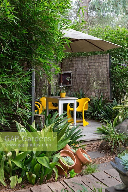 Terrasse en bois recouverte d'un grand parapluie blanc avec des chaises rétro en plastique jaune vif et une table en bois, recouverte de stores allumettes en bambou et encadrée par une touffe de bambou et Aspidistra elatior, usine de fonte.