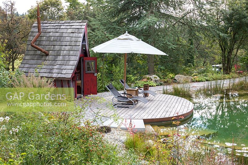 Terrasse en bois ronde avec chaises en bois et parasol devant le chalet des sorcières. Les plantes comprennent Calamagrostis brachytricha, Cedrus libani