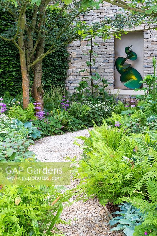 Le jardin Morgan Stanley - chemin de gravier à travers le côté du jardin menant à la sculpture en alcôve dans le mur de pierres sèches, sculpture de Craig Schaffer. La plantation comprend Primula beesiana, hostas et fougère royale.