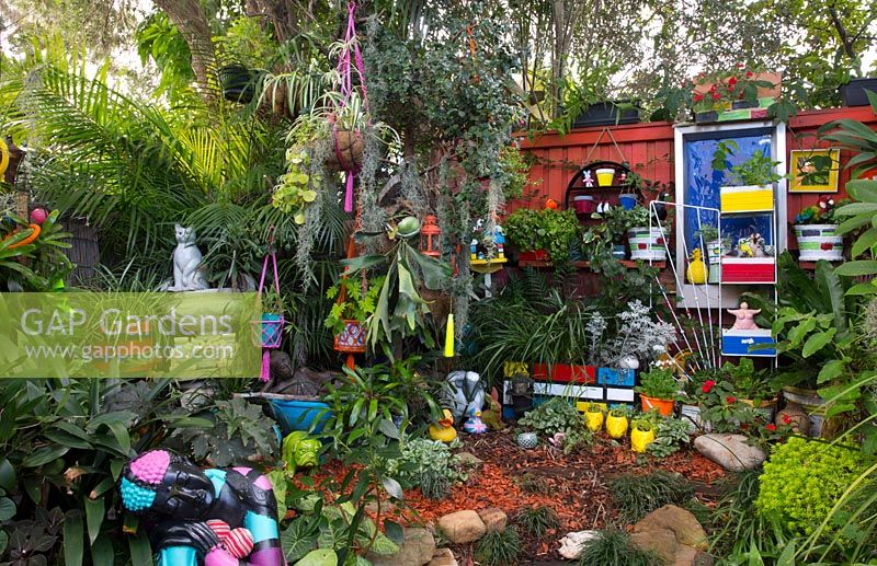 Zone arrière surélevée du jardin avec une collection éclectique d'ornements de jardin, de pots, de statues et de paniers suspendus parmi des plantes aimant l'ombre.