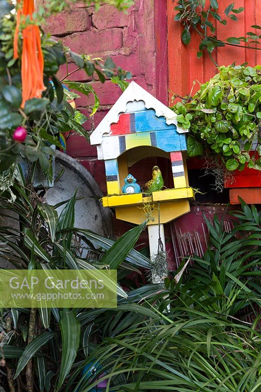 Une maison d'oiseau ornementale d'inspiration mondrienne parmi des plantes à feuilles lanières appuyées contre un mur rose et orange.
