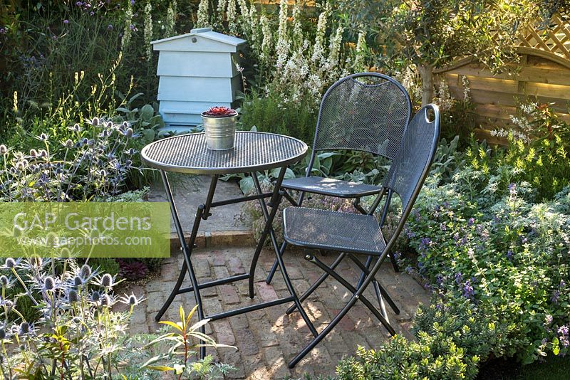Petit jardin de style chalet d'été avec terrasse en brique, table et chaises en métal et plantation en argent blanc et bleu.