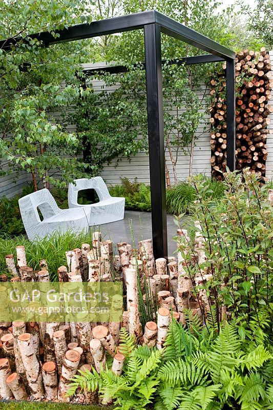 Le jardin naturellement à la mode conçu par NDG. Coin salon avec sièges en maille métallique, pergola noire, rondins de bouleau
