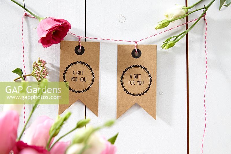 Étiquettes-cadeaux artisanales de la Saint-Valentin suspendues à une guirlande de ficelles avec des fleurs coupées.