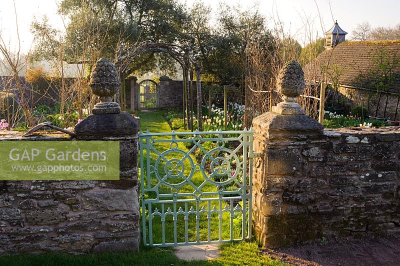Porte d'accès au jardin de coupe où un chemin d'herbe mène sous quatre poutres blanches dressées, Sorbus aria 'Lutescens', vers une porte d'accès au potager. Ferme Brilley Court, Whitney-on-Wye, Herefordshire