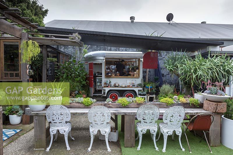 Vue d'un café en plein air, avec un bar flottant pour chevaux converti et une longue table en bois avec jardinières intégrées et chaises en fonte blanche, Australie.